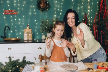 Anne ve kızı süslenmiş bir mutfakta Noel kurabiyeleri hazırlarken kurabiye kesicileri gösteriyor.