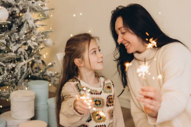 Anne ve kızı birbirlerine havai fişek tutuyorlar ve Noel ağacının yanında hediye kutularıyla gülümsüyorlar.