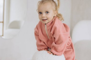 Little girl with blond hair and pink fleece jumpsuit is posing in a bright room for clothing mockup clipart