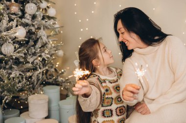 Anne ve kızı havai fişek tutuyor, Noel arifesini süslenmiş ağacın ve hediye kutularının yanında kutluyorlar, birbirlerine bakıyorlar.