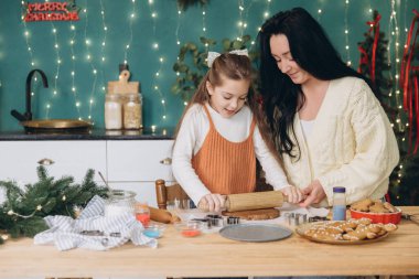 Anne ve kızı neşeli bir şekilde hamuru sarıyorlar ve Noel neşesiyle dolu güzel dekore edilmiş bir mutfakta Noel kurabiyeleri yapıyorlar.