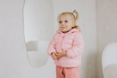 Beyaz odada aynanın yanında duran pembe ceketli ve pantolonlu gülümseyen sarışın kız.