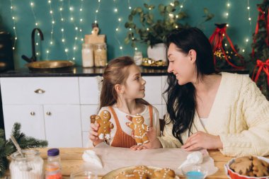 Anne ve kız birbirine bakıyor, zencefilli kurabiye gösteriyor Noel tatilinde, dekore edilmiş bir mutfakta