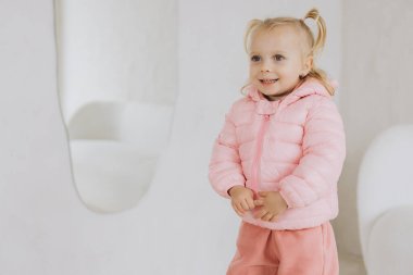 İki at kuyruklu, pembe ceketli ve pantolonlu, parlak bir odada beyaz mobilyalarla poz veren sevimli küçük bir kızın portresi.