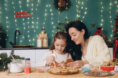 Anne ve kızı Noel için dekore edilmiş bir mutfakta zencefilli kurabiye süslüyor.