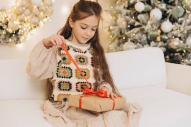 Gülümseyen kız, Noel hediyesinde kırmızı kurdeleyi çözerken süslü ağaç ve bokeh ışıklarının yanındaki kanepede oturuyor.