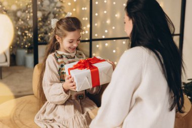Mutlu kız annesinden noel hediyesi alıyor, aydınlanmış noel ağacının yanında oturuyor, evde kış tatilinin tadını çıkarıyor.