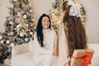 Arkasında Noel hediyesi saklayan bir kız annesine sürpriz yapıp Noel ağacının yanındaki koltukta oturuyor.