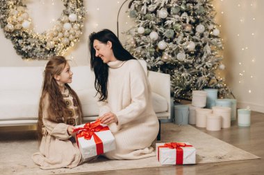 Anne ve kızı Noel hediyelerini değiş tokuş ediyor Noel ağacının yanında yerde oturuyor, rahat kışlık giysiler giyiyorlar.