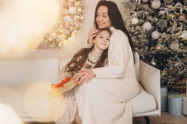 Anne ve kızı bir kanepede Noel süslemeleri ve arka planda bir noel ağacıyla sarılıp bir Noel hediyesini paylaşıyorlar.