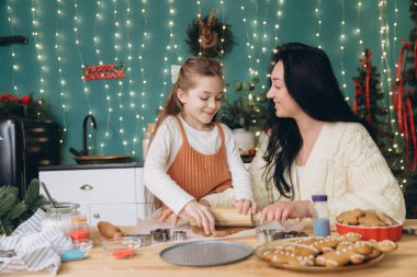 Anne ve kız hamuru yuvarlıyor ve kurabiye keskisi kullanıyor, yılbaşı için mutfakta zencefilli kurabiye pişiriyor, ışık ve süslemelerle süslenmiş.