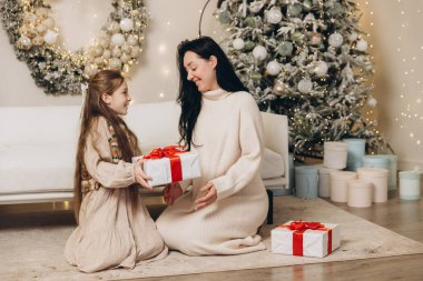 Güzel dekore edilmiş bir ağacın yanında oturmuş, neşeli bir şekilde annesine Noel hediyesi veriyor.
