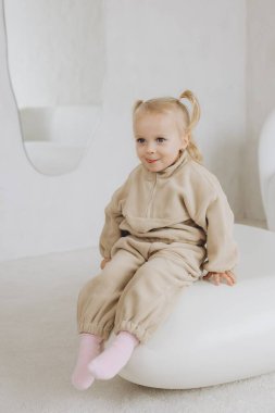 İki at kuyruklu, bej eşofmanlı, pembe çoraplı parlak odada beyaz bir puf üzerinde oturan küçük kız.