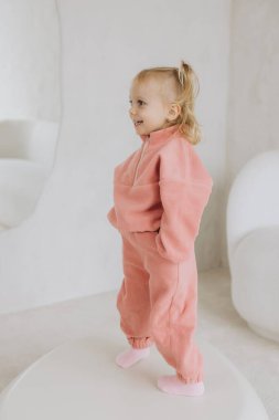 Süslü, pembe eşofmanlı, yuvarlak beyaz bir masa üzerinde modern bir iç mekânda duran sevimli, küçük kız.