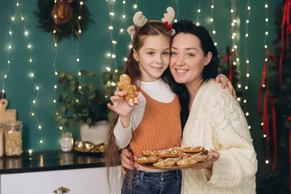 Anne ve kızı Noel için dekore edilmiş mutfakta zencefilli ekmek ve kurabiye tutuyor ve gösteriyorlar.