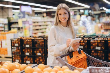 Genç bir kadın markette alışveriş yaparken not defterine bir şeyler yazıyor. Sağlıklı listesi için eşyaları dikkatle kontrol ediyor.