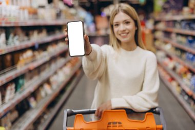 Süpermarkette alışveriş yaparken beyaz ekranlı akıllı telefon gösteren gülümseyen bir müşteri