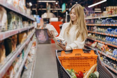 Genç bir kadın süpermarkette alışveriş yaparken iki farklı gıda ürününü bir alışveriş arabasıyla karşılaştırıyor.