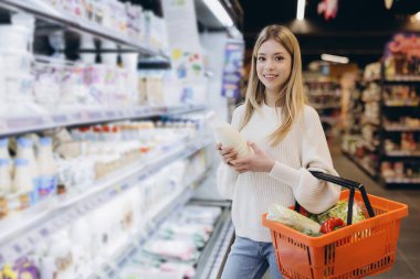 Elinde süt şişesi ve alışveriş sepeti olan genç bir kadın süpermarket süt ürünleri reyonundan alışveriş yapıyor.