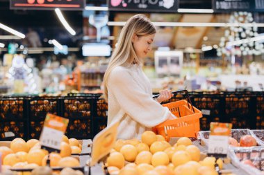 Genç bir kadın taze ürün alışverişi yapıyor, elinde turuncu bir sepet tutuyor ve modern bir süpermarketin meyve ve sebze reyonunda greyfurt seçiyor.