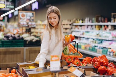 Genç bir kadın taze sebze alışverişi yapıyor, elinde dolmalık biber tutuyor ve marketten çeşitli ürünler seçiyor.