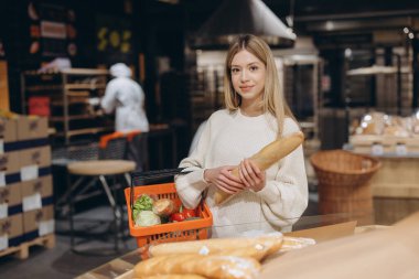Bakkal alışverişi yapan genç bir kadın, elinde bir baget ve bir sepet dolusu sebzeyle bir süpermarketin pastanesinde