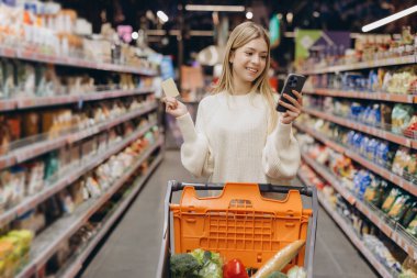 Marketten alışveriş yaparken akıllı telefon kullanan ve kredi kartı tutan genç bir kadın.