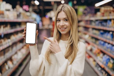 Marketteki boş akıllı telefon ekranına işaret eden genç bir kadın, alışveriş için mobil uygulama öneriyor