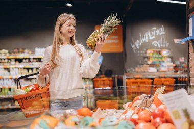 Parlak, modern bir süpermarketin üretim bölümünde alışveriş yaparken elinde olgun bir ananas tutan genç bir kadın, gülümsüyor ve taze seçenekleri inceliyor.