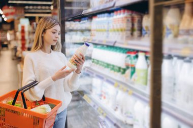 Alışveriş sepeti tutan ve süt şişesini marketin dondurulmuş süt bölümünde seçen genç bir kadın.