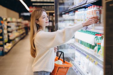 Genç bir kadın market alışverişi yapıyor. Parlak ışıklı bir süpermarkette soğutulmuş raflardan bir şişe süt alıyor.