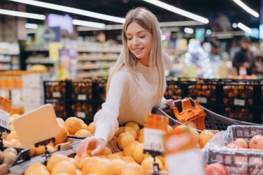 Genç bir kadın süpermarketten portakal alıyor, raftan meyve topluyor ve alışveriş arabasına koyuyor.