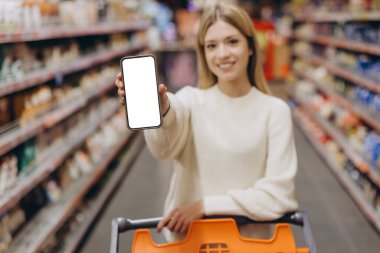 Süpermarkette alışveriş yaparken beyaz ekranlı akıllı telefon gösteren gülümseyen bir müşteri