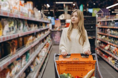 Markette alışveriş arabalarını iten genç bir kadın raflardan yiyecek ürünleri seçiyor, market alışverişi yapıyor.