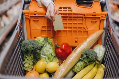 Bir süpermarkette içi taze ürünlerle dolu bir alışveriş arabasının üzerinde kredi kartı tutuyor.