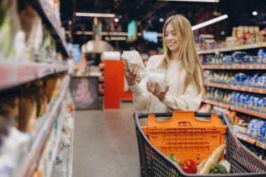Gülümseyen kadın, açık, düzenli bir süpermarkette alışveriş yaparken iki market malzemesini karşılaştırıyor, taze ürün seçme deneyiminin tadını çıkarıyor.