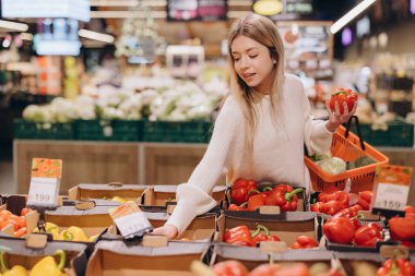 Genç bir kadın taze ürün alışverişi yapıyor, parlak ışıklı bir süpermarkette sergiden biber topluyor.