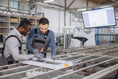 İki fabrika işçisi alüminyum pencere çerçevesini bir üretim hattında birleştiriyor, dijital ekrandaki talimatları kontrol ediyorlar.