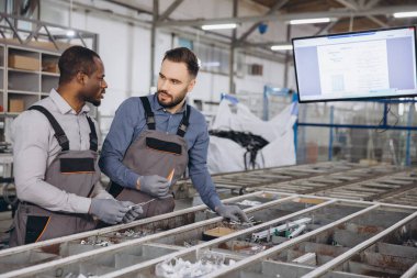 Tulumlu ve eldivenli iki fabrika işçisi, taşıyıcı bandın yakınındaki alüminyum pencere ve kapıların üretim sürecini tartışıyor.