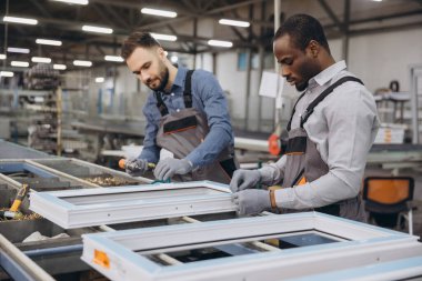 İki fabrika işçisi PVC pencere çerçevelerini bir üretim hattında birleştiriyor, yoğun bir endüstriyel ortamda takım çalışması ve beceri gösteriyor