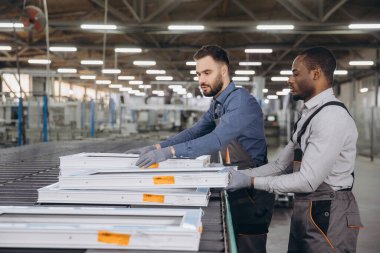 İki fabrika işçisi PVC pencere çerçevelerini modern bir üretim tesisinde bir taşıyıcı bandın üzerine dikkatlice yerleştiriyor.