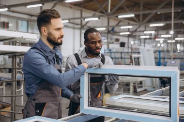 Modern bir imalat tesisinde kalite kontrolü sırasında yeni üretilen bir pvc pencere çerçevesini dikkatle inceleyen iki fabrika işçisi