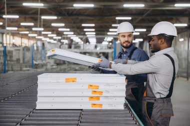 Güvenlik kaskı ve eldiven takan iki fabrika işçisi modern bir fabrikadaki bir üretim bandında PVC pencere çerçeveleri taşıyorlar.