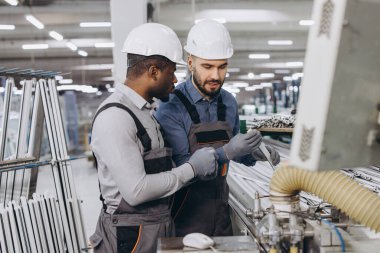 Kasketli ve tulumlu iki fabrika işçisi modern bir pencere ve kapı üretim tesisinin yakınındaki bir makinenin alüminyum profillerini kontrol ediyorlar.