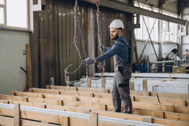 Koruyucu iş kıyafeti ve kask giyen fabrika işçisi pencere ve kapı yapımı için alüminyum profilleri kaldıran bir vinç işletiyor.