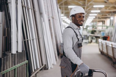 Gülümseyen Afro-Amerikan fabrika işçisi alüminyum ve PVC pencere ve kapı profillerini büyük bir depoda el arabası kullanarak taşıyor.