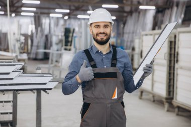 Üniformalı, kasklı, alüminyum pencere profilli gülümseyen fabrika işçisi atölyede başparmak hareketi yapıyor.