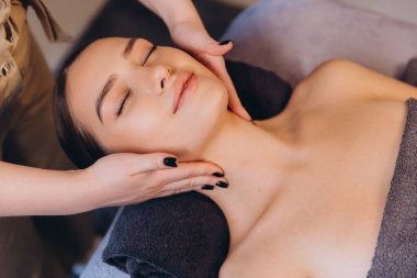 Güzellik uzmanı genç bir kadının yüzüne ve boynuna rahatlatıcı bir masaj yapıyor. Huzurlu bir spa salonundaki masaj masasında rahatça yatıyor.