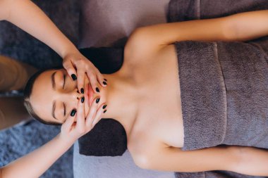 Masöz masaj salonundaki masaj masasında uzanan genç bir kadına yüz masajı yapıyor.