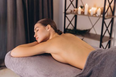 Masaj masasında uzanan rahat bir kadın, arka planda mumlarla masaj terapisinin tadını çıkarıyor.